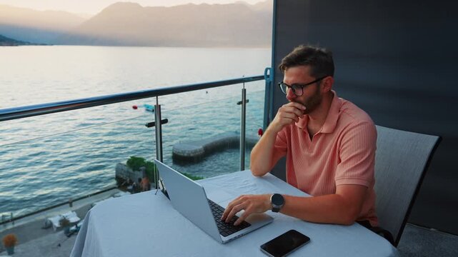 Remote worker typing on laptop at sunset balcony overlooking calm sea and mountains, enjoying flexible lifestyle with outdoor workspace and scenic water view