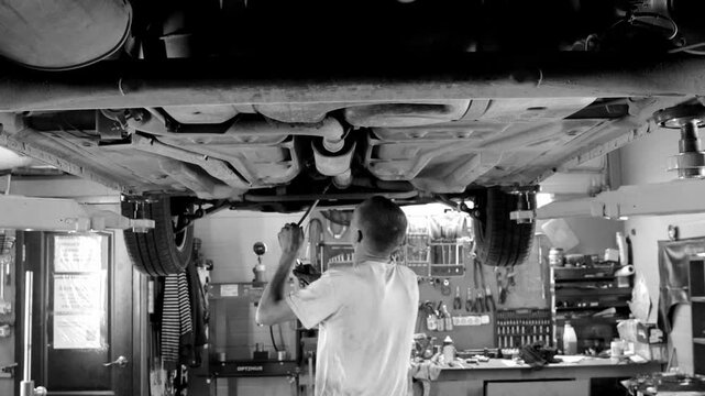 mechanic inspecting the underside of a raised car in a busy auto repair garage with tools, equipment, and workshop details visible black white video