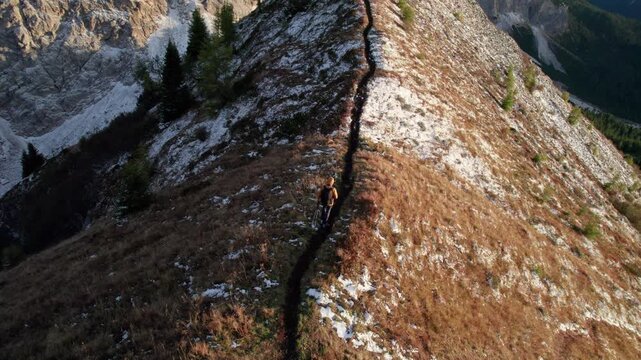 Top-down drone view follows solo hiker with backpack and trekking poles along narrow mountain ridge trail, surrounded by patchy snow and autumn alpine vegetation in Dolomites, Italy