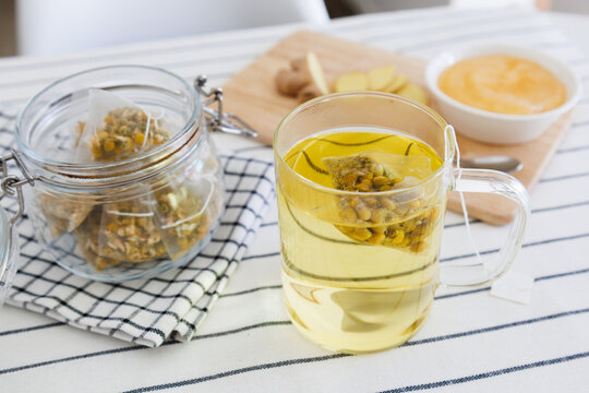 A glass cup of chamomile tea with a tea bag, honey, and sliced ginger on a kitchen table.
