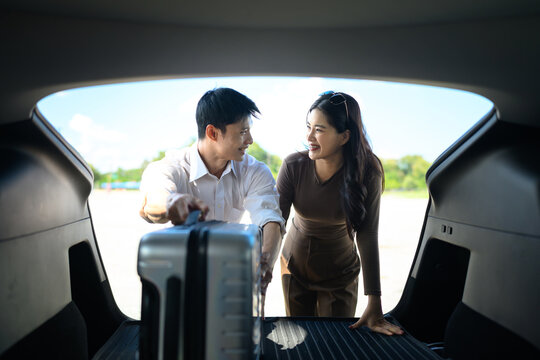 Happy Asian couple loading suitcase into car trunk, smiling at each other before road trip vacation