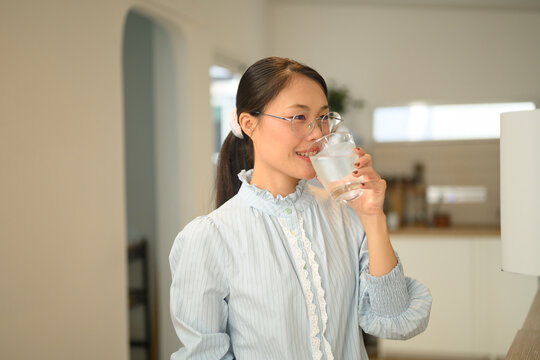 Happy Asian woman drinking a glass of water at home in a bright domestic kitchen
