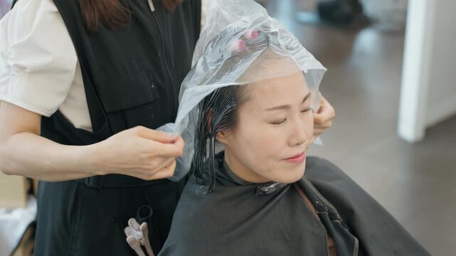 Hairdresser in Korean beauty salon covers female client hair rolled in perm rods with plastic wrap to lock in heat and moisture during permanent wave treatment