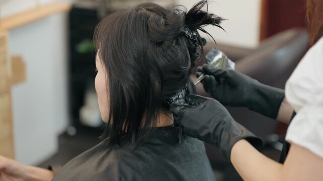 Hairdresser wearing black gloves holds section of client hair and applies chemical solution with brush while remaining hair is clipped up in modern beauty salon