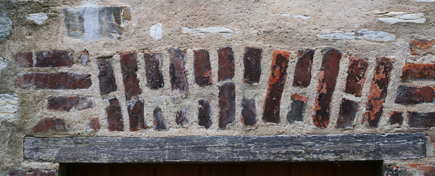 rustic, weathered brick lintel or segmental arch set into a rough lime mortar wall, positioned directly above a thick, aged horizontal wooden beam that forms the top frame of a doorway