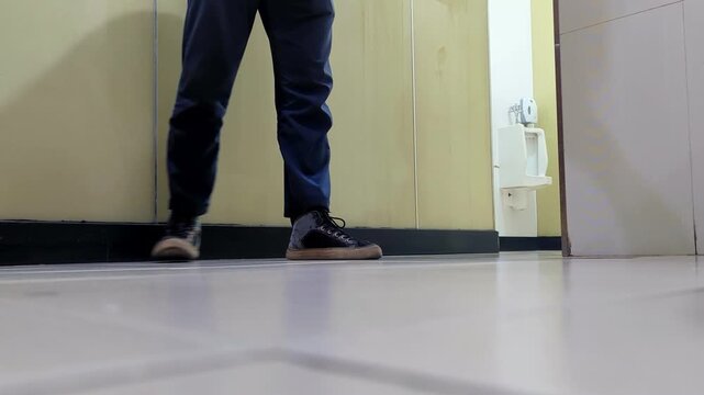 A janitor mops the tiled floor of a office restroom, wearing dark trousers and sneakers while cleaning near a wall-mounted urinal.
