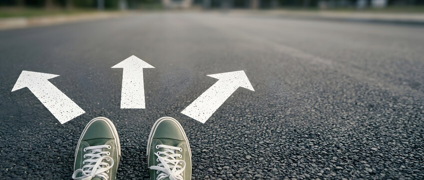 A first-person perspective of a person wearing green sneakers standing on an asphalt road at a crossroads with three white arrows pointing in different directions, symbolizing making life choices.