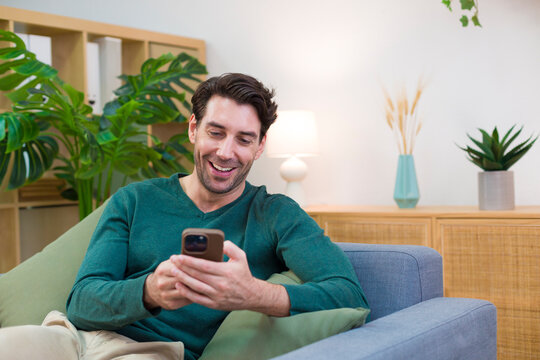 White man sitting on sofa smiling using smartphone in living room