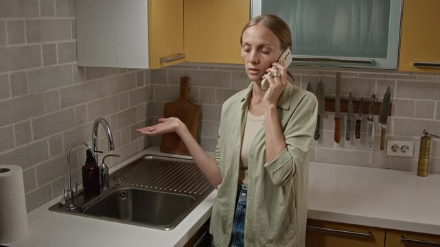 Young adult Caucasian woman talking on mobile phone and gesturing near broken water faucet in kitchen. Clip useful for plumbing emergency services and home repair commercials