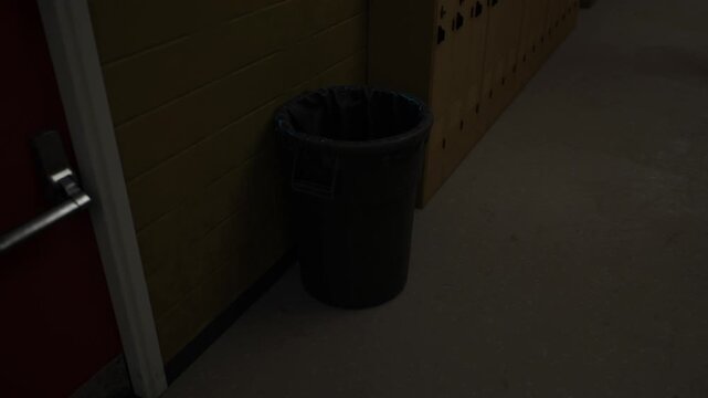 In a quiet hallway, a black trash can stands against the wall. The subdued lighting creates a sense of emptiness, highlighting the cans presence in the otherwise bare space.