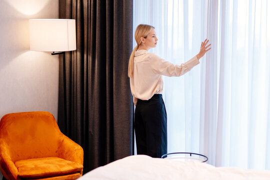 Woman beside a bedroom window adjusting curtains for home comfort. She turns to the camera with her hand raised to sheer fabric, showing a domestic moment framed by the bed and softly lit window.