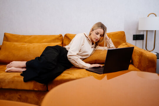 Woman reclining on a sofa with her laptop at home in the evening. She rests her head on one hand and uses the touchpad with the other, keeping the computer beside her during a break after work.