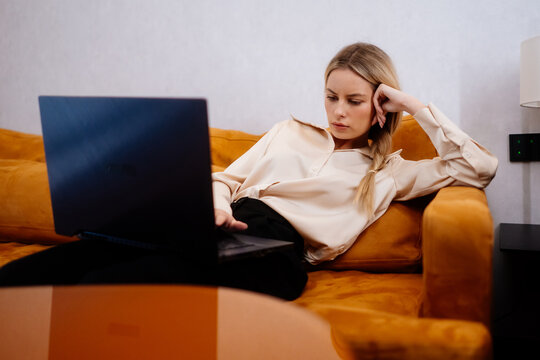 Tired businesswoman resting with a laptop on the sofa at home. She reclines against the cushions with one hand supporting her head, looking at the computer during a quiet evening after work.