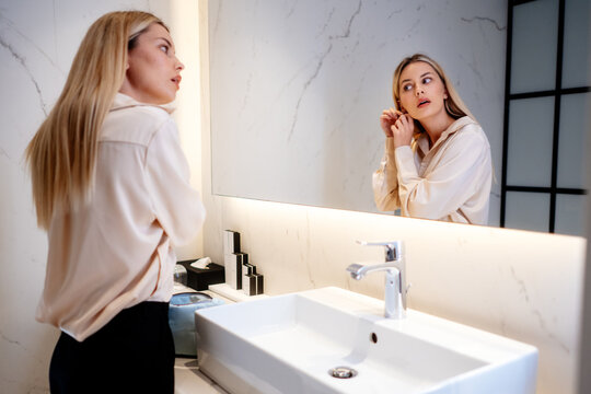 Woman accessorizing her outfit with earrings at a bathroom mirror. The wider view includes the washbasin as she raises her hands to her ear and checks the accessory in her reflection.