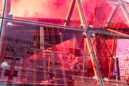 Bright red translucent glazing and metal joints reflecting neighboring buildings in a modern architectural detail with overlapping surfaces and vivid warm light