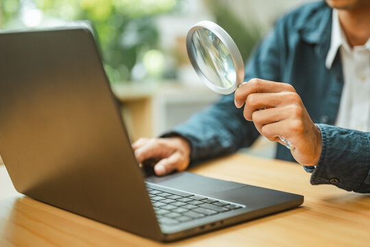 Online research with a magnifying glass held toward a laptop; inspection of computer information represented by a lens and careful inspection at an indoor desk without readable screen details.