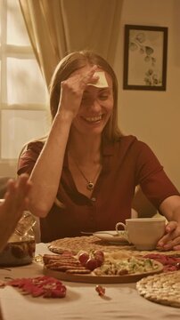 Vertical video of Caucasian young adult woman playing forehead sticky note guessing game at table. Suitable for lifestyle advertising, board game marketing, and social connection campaigns