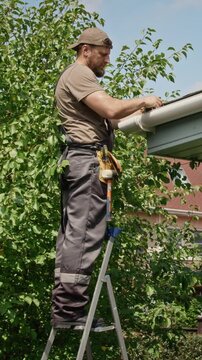 Vertical video of Caucasian young adult worker standing on ladder, removing debris from roof gutter. Clip suitable for home improvement advertising and property maintenance campaigns