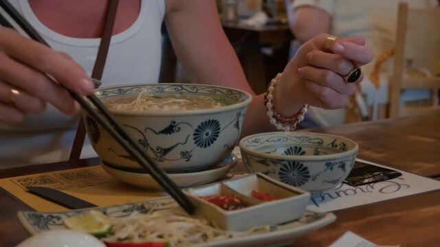 Person using chopsticks to lift noodles from patterned ceramic bowl during casual restaurant meal at wooden table with condiments and garnishes visible on tabletop