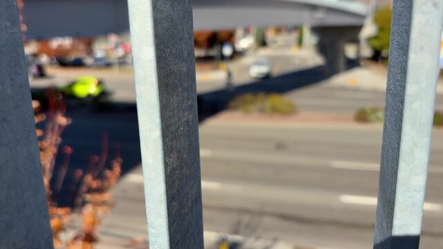 Soft-focus of traffic moving on a busy two-way road on a sunny day, through the metal railings of a pedestrian overpass. 