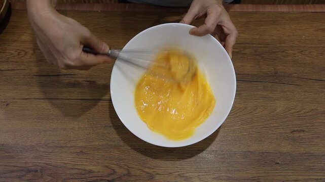A cook uses a whisk to beat eggs together a moderate speed in a white bowl on a wood-grain table to prepare dishes such as omelets, silky steamed eggs, or scrambled eggs. Crack eggs into a bowl
