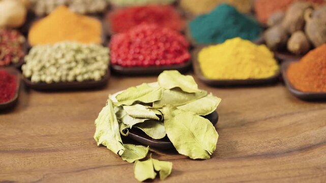 aromatic dried kaffir lime leaves in a square bowl on a wooden table, surrounded by various spices and herbs.