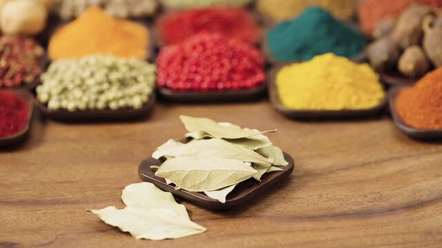 fragrant dried bay laurel leaves in a square bowl on a wooden table, surrounded by various spices and herbs.