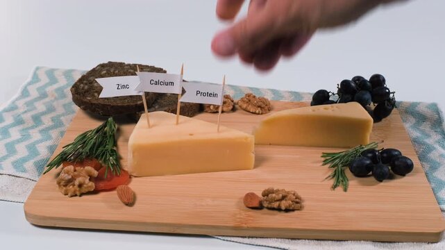 A hand inserts zinc, phosphorus, protein, calcium and vitamin A flags into cheese beside bread, grapes and nuts. Beneficial dairy nutrients and varied eating concept