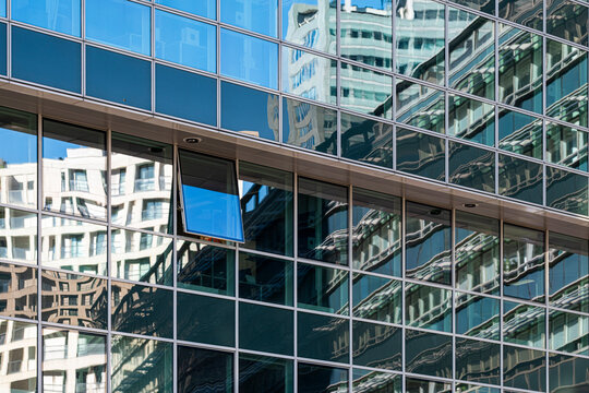 Blue glass facade with a regular window grid and reflections of neighboring modern buildings and layered urban architecture in a geometric composition