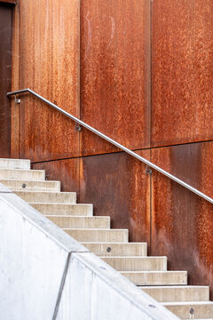 Concrete stairs with a diagonal metal handrail alongside a rust colored steel wall showing material contrast in modern exterior architecture