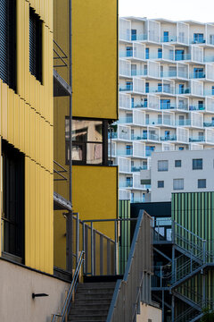 Yellow building with an exterior staircase leading toward a blue and white apartment facade with repeated balconies in a narrow urban courtyard