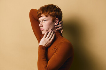 Young man with red hair posing with hand on neck in brown turtleneck sweater against beige background. Side profile portrait of serious confident male model in soft studio light. © SHOTPRIME STUDIO