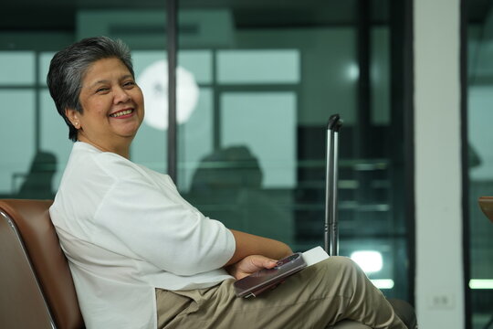 An older Thai woman travelling alone laughs at a message on her phone on the seats of an airport gate area, with her wheeled case beside her, an authentic look at ageing and technology