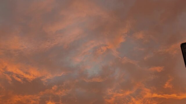 Orange illuminated clouds slowly shift across a moody evening sky, with a dark building edge visible along the right side of the frame