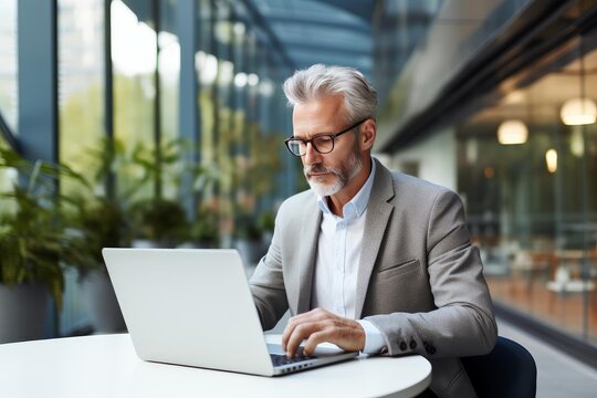 Mature businessman with gray hair and beard wearing glasses, focused on working remotely at a laptop, demonstrating dedication and productivity in a modern office or coworking space