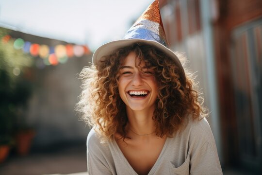 Joyful young woman with curly hair wearing a festive party hat, laughing out loud, celebrating a happy occasion outdoors under bright sunlight, feeling excited and cheerful