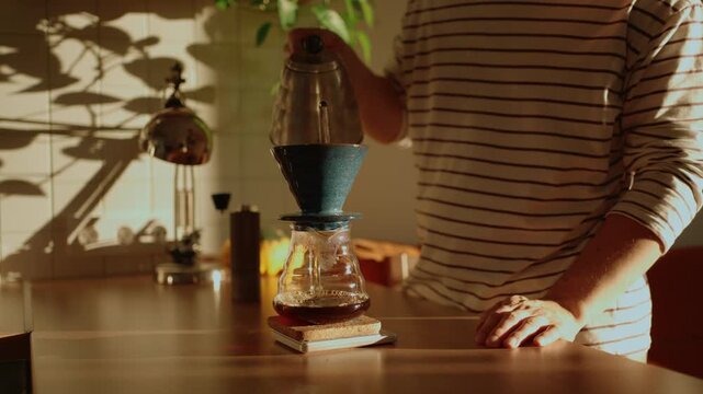 Man making specialty pour over coffee in a sunlit kitchen, carefully pouring hot water from a gooseneck kettle over grounds in a dripper to brew a fresh cup for his morning routine