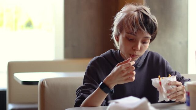 In this lively video, a teenage girl with vivid hair eats French fries at a fast-food cafe, portraying a relaxed everyday experience and the joy of enjoying a favorite snack.