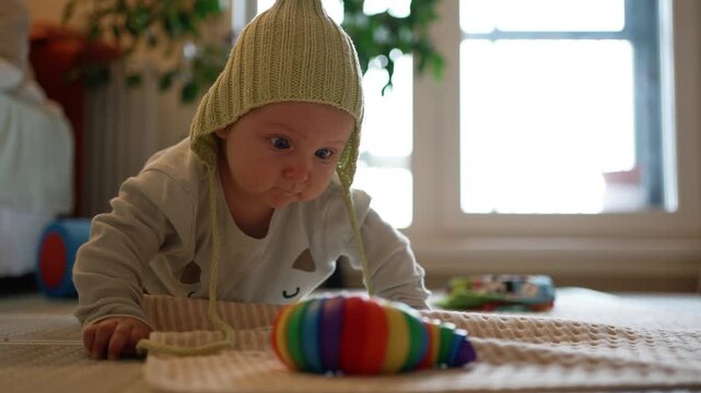 Adorable little baby in a green pixie hat lying on his stomach and playing with a bright rainbow toy on the floor. Cute newborn discovering the world and learning through play