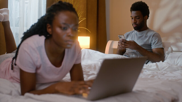 African American woman browsing internet typing laptop in bed boyfriend chatting mobile phone relaxed couple ethnic family weekend online computer work gadget addiction indoor home bedroom evening