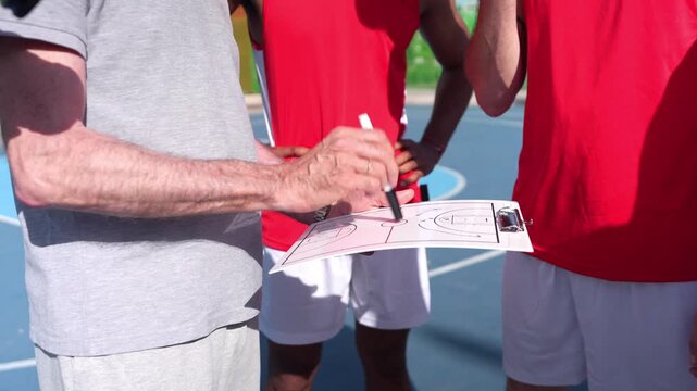 Closeup of a senior coach drawing a game plan on a clipboard, explaining the basketball tactic to a diverse team of players on an outdoor court
