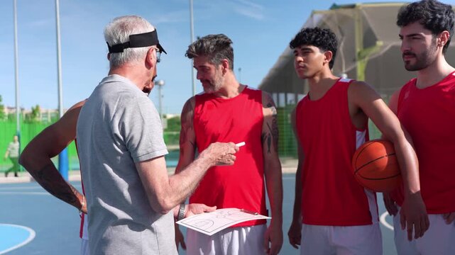 Senior male coach discussing a new tactic with a diverse team of players on an outdoor basketball court. Teamwork and leadership concept