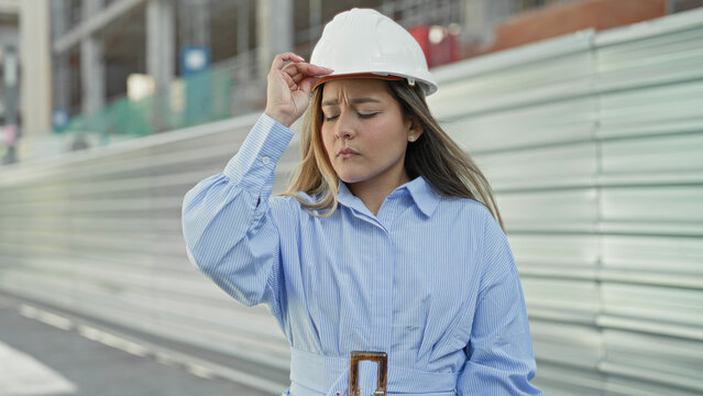 Woman architect in white hardhat adjusts helmet while touching her hair at a building construction site by corrugated metal hoarding; determination focus.