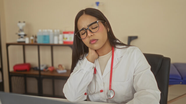 Young woman doctor rubbing neck with stethoscope at desk in building, eyes closed and laptop visible, shelf with medical supplies behind; fatigue telemedicine.