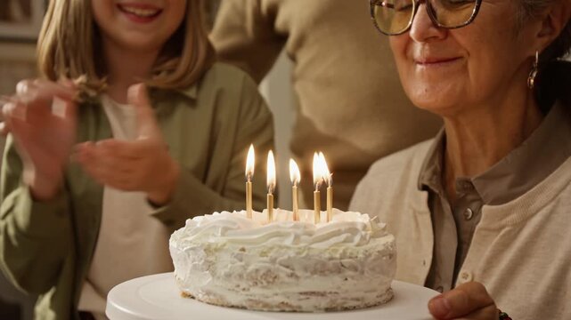 Senior Caucasian woman blowing out candles on birthday cake while Caucasian child claps, close up. Useful for elderly care advertising and family lifestyle campaigns
