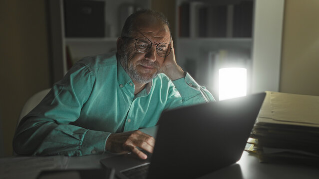 Man using laptop, resting head on hand while typing with one hand in a dim office building at night; late night fatigue.