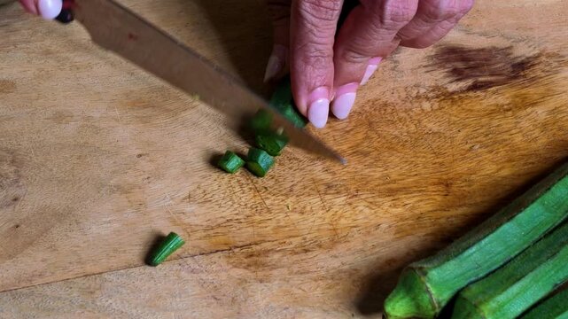 Closeup of Indian woman's hands cutting fresh green okra or bhindi with a kitchen knife on a wooden chopping board, preparing vegetables for traditional Indian cooking and homemade meals.
