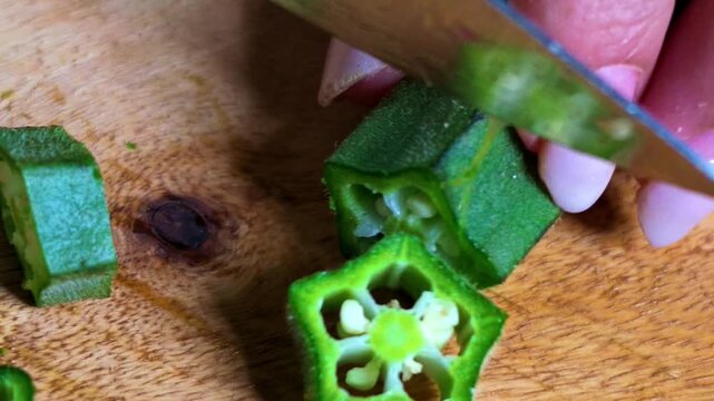 Closeup of Indian woman's hands cutting fresh green okra or bhindi with a kitchen knife on a wooden chopping board, preparing vegetables for traditional Indian cooking and homemade meals.