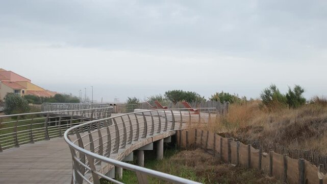 Modern pedestrian promenade, Mediterranean resort. Terracotta lounge chairs, villa roofs, and dune grasses. Muted pastel colors and a hazy white sky. Hospitality and tourism industry.