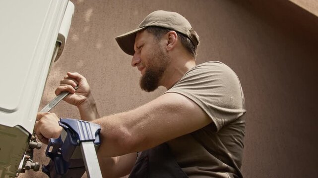 Middle aged Caucasian technician standing on ladder, using wrench to install outdoor air conditioner unit. Ideal for HVAC service advertising and home repair promotions
